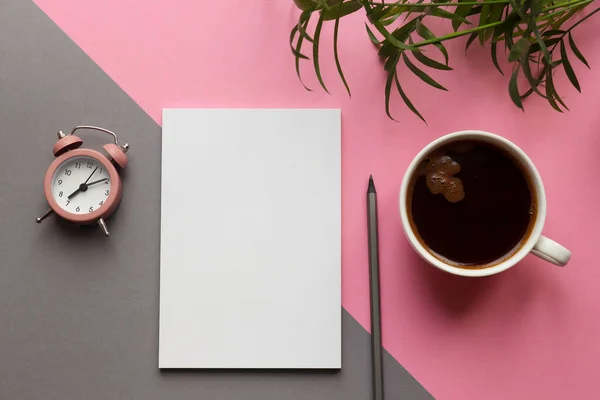 Morning concept, start of work. Modern workplace with notepad, pencil, plant, cup of coffee and alarm clock on the pink and grey desk. Business and education flat lay. Top view, copy space.