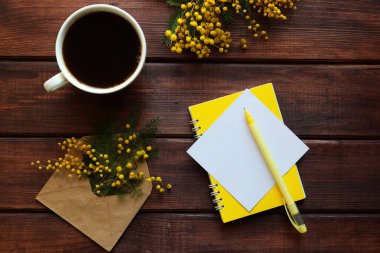Bir fincan kahve, mimoza çiçekleri, sarı not defteri, boş kağıt ve tahta arka planda bir kalem. Mimoza çiçekleri ile kırsal bahar arka planı. Üst manzara, düz uzanma.
