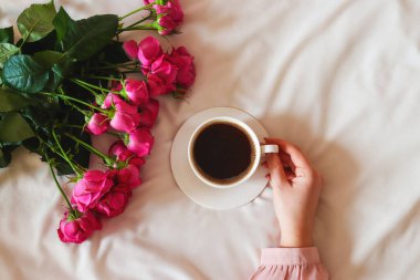 Bir kadının eli yatakta bir fincan kahve tutuyor. Rahat bir sabah bezi, çiçekler ve bir fincan kahveyle. Boşluğu kopyala, minimalizm, üst görünüm.