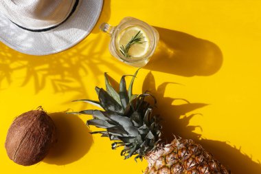 Parlak yaz kompozisyonu. Hindistan cevizi, ananas, soğuk limonata ve sarı arka planda koyu gölgeli bir şapka. Yaz konsepti, tatil konsepti. Üst manzara, düz uzanma.