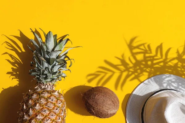 Parlak yaz kompozisyonu. Hindistan cevizi, ananas ve sarı arka planda koyu gölgeli bir şapka. Yaz, tatil konsepti. Üst manzara, düz uzanma.