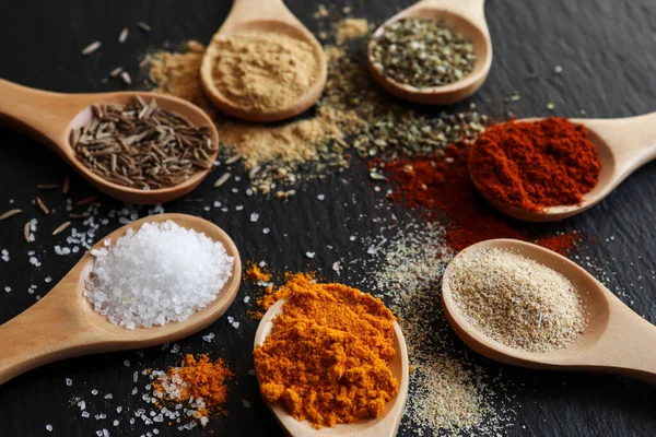 Koyu renkli bir masa üzerinde ahşap kaşıklarda çeşitli baharatlar ve otlar. Paprika, zerdeçal, granüllü sarımsak, kurutulmuş zencefil, kimyon, kurutulmuş otlar ve deniz tuzu. Seçici odak.