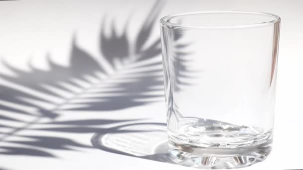 Czysta woda wlewa się do szklanki na stole z cieniem liści palmowych. Koncepcja detoksykacji i letnich napojów orzeźwiających. Zbliżenie — Wideo stockowe