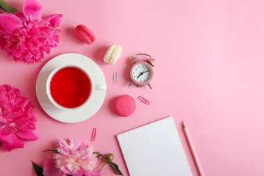 Not defteri, kalem, çalar saat, şakayık çiçekleri, açık pembe arka planda bir fincan çay ve badem kurabiyesi ile yatıyordu. İş, serbest meslek ve eğitim kavramı. Sabah oldu. Üst görünüm.