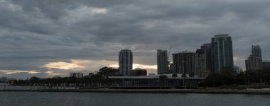 St. Petersburg silueti, Tampa Körfezi 'nin rıhtımındaki bulutların üzerinden yumuşak bir ışık geçerken dramatik bir akşam gökyüzüne karşı siluette belirir. Yüksek binalar ve şehir merkezindeki kuleler tanınabilir bir Körfez Kıyısı profili oluşturuyor..