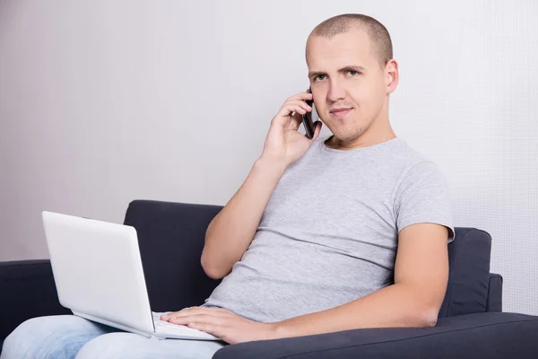 Jeune bel homme assis sur le canapé avec ordinateur et mobile phon — Photo