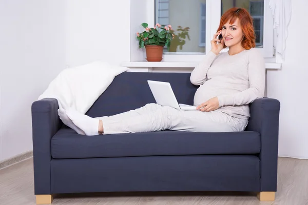 Молода вагітна жінка використовує ноутбук і розмовляє по телефону вдома — стокове фото