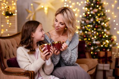 Noel ve yeni yıl konsepti. Annesiyle çay ya da sıcak çikolata içen tatlı bir kız. Noel ağacı ve tatil ışıklarıyla süslü oturma odasında.