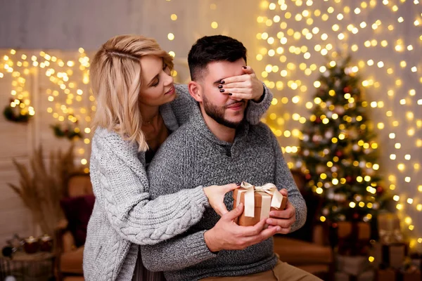 Christmas, New Year, love and surprise concept - beautiful blonde woman giving Christmas present to her boyfriend in decorated living room with Christmas tree and lights