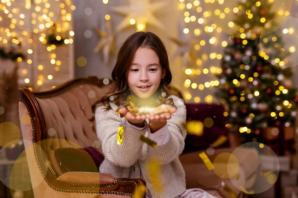 Christmas, childhood and magic concept - portrait cute little girl blowing fairy dust off her palms in decorated living room with Christmas tree and holiday lights