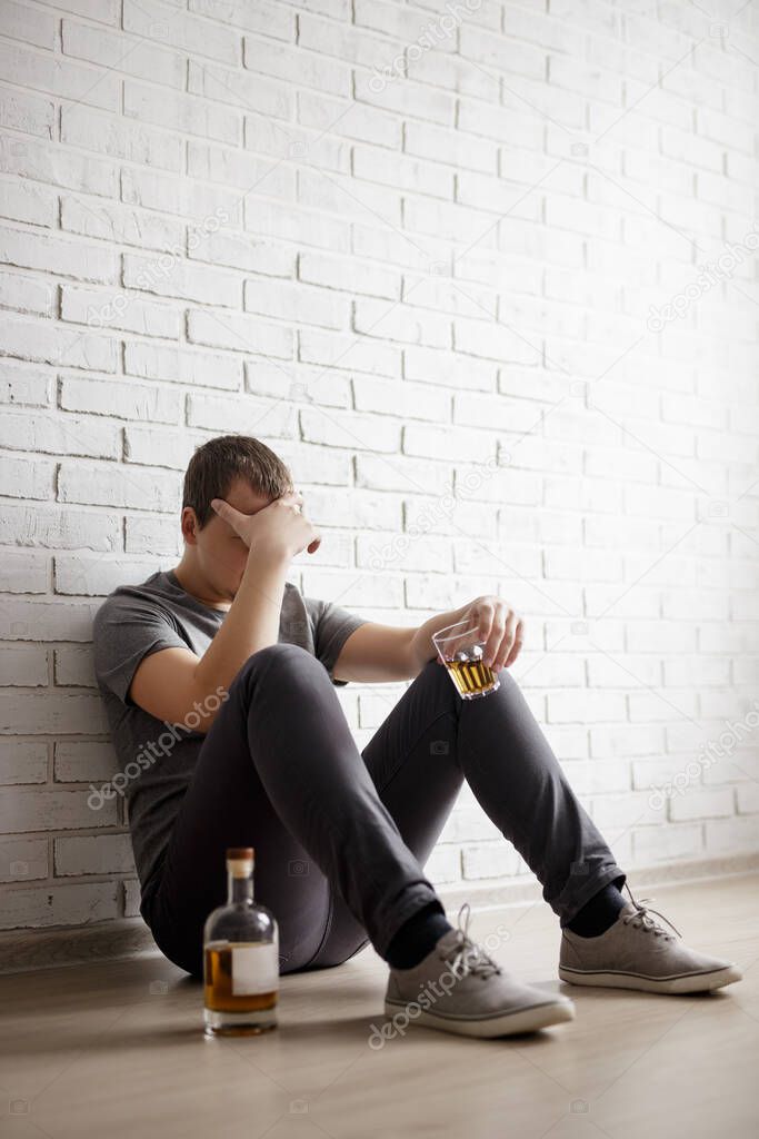 young man suffering from alcoholism, guy sitting on the floor with glass and bottle of whiskey