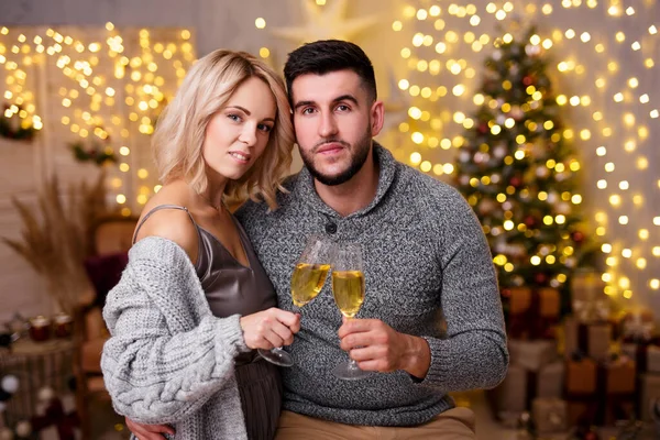 Noel 'i ya da yeni yılı kutlayan genç ve güzel bir çift Noel ağacı, hediye kutuları ve ışıkları olan dekore edilmiş oturma odasında şampanya içiyorlar.