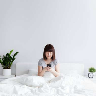 Mutlu kadın yatakta oturuyor ve evde akıllı telefon kullanıyor. Beyaz duvar arkaplanının üzerine boşluğu kopyalıyor.