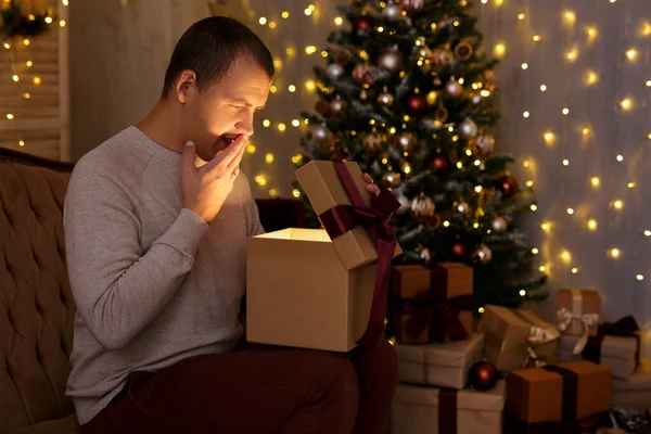 Christmas and magic concept - portrait of surprised young man opening gift box in decorated dark room with Christmas tree and holiday led lights