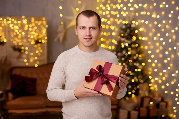 冬の休日のコンセプト クリスマスツリーとLedライトと装飾されたリビングルームでギフトボックスを持つ若いハンサムな男 — ストック写真
