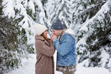 Genç romantik çiftin portresi kış ormanında el ele tutuşup soğukta ısınmak için esiyor.