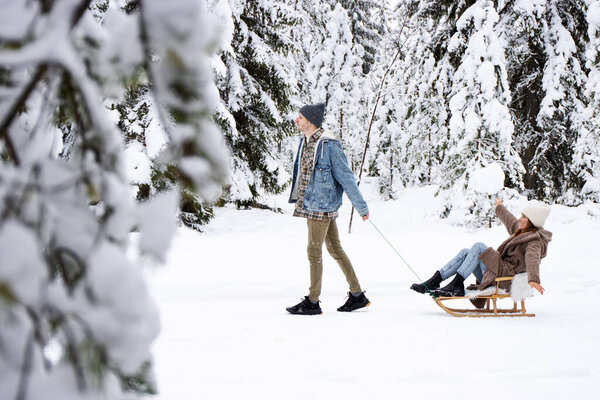 man pulling sledge with joyful woman on winter day in snow forest
