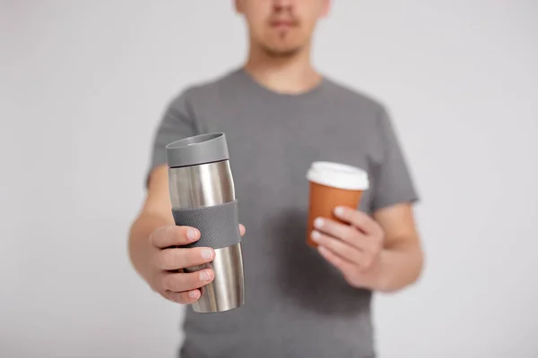 zero waste and eco friendly concept - thermo cup and disposable paper coffee cup in male hands over grey background with copy space