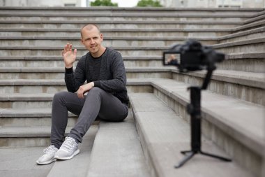 Genç yakışıklı blog yazarı beton merdivenlerde oturuyor ve modern DSR kamerasıyla video kaydediyor.