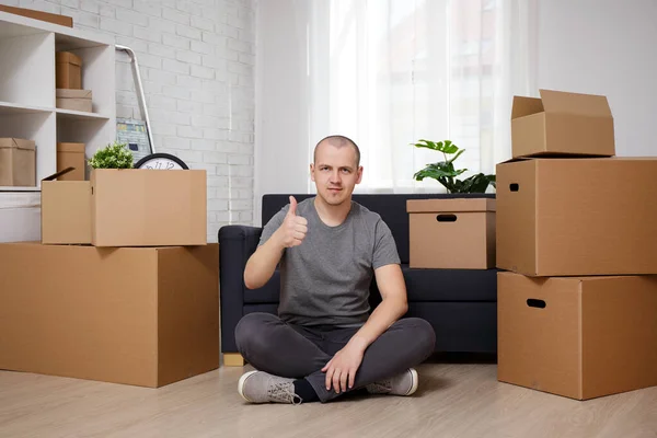 moving day - hadsome man sitting on floor in his apartment with moving boxes and thumbing up