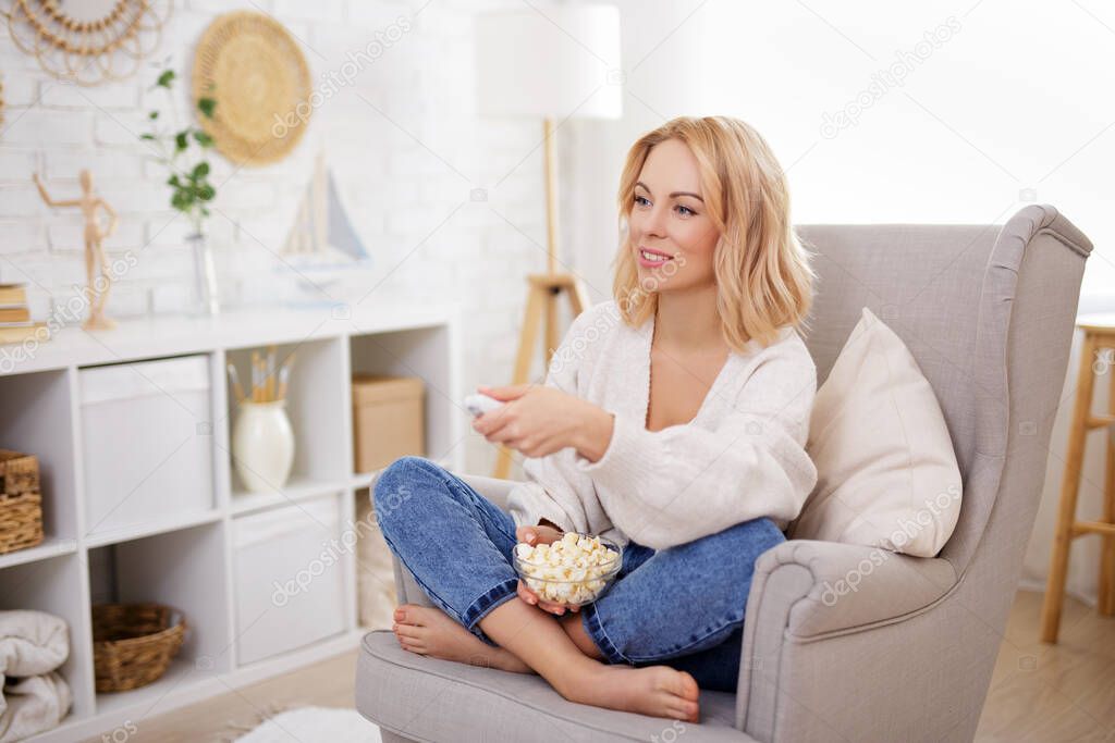 happy young beautiful woman eating popcorn, watching tv and changing channels with remote control in bright living room