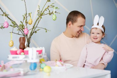 Aile, mutluluk ve bahar tatili konsepti. Mutlu baba portresi. Küçük tatlı kızı ve kremalı kekler ve renkli paskalya yumurtalarıyla süslenmiş masa.