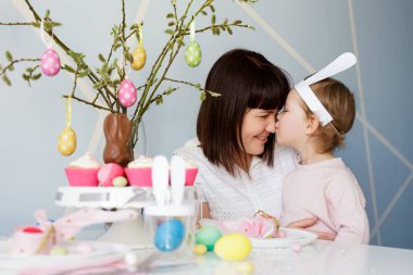 Family and Easter concept - happy mother with cute little daughter and decorated table pussy willow branch with cupcakes, colorful painted Easter eggs and decorations