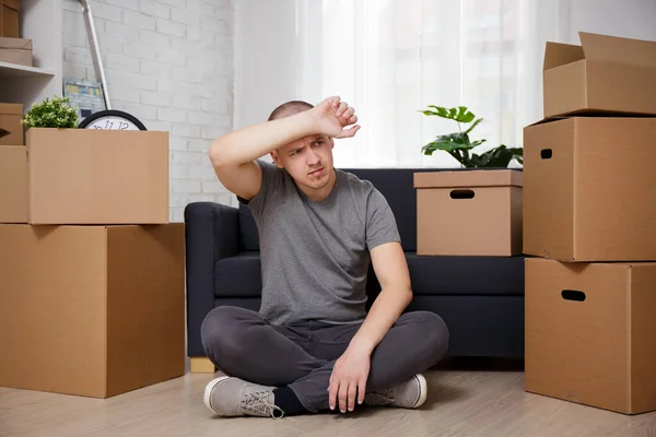 moving to new home - tired man sitting on floor in his new apartment with cardboard boxes after moving day