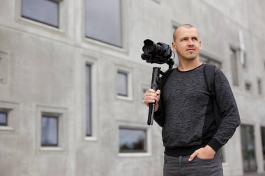 Profesyonel erkek videografın portresi DSR kamerayla 3 eksenli dengeleyici üzerinde poz vermek, gri beton bina üzerine kopyalama alanı