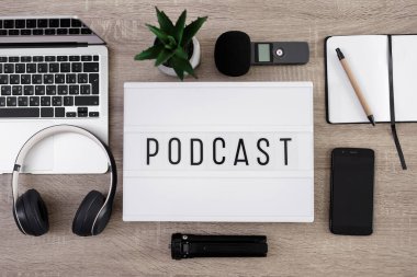 Podcast ve blog konsepti - ses kayıt ekipmanlarıyla düz zemin, dizüstü bilgisayarı, mikrofonu, kulaklığı, akıllı telefonu ve not defteriyle birlikte tahta masanın üzerinde podcast sözcüğü