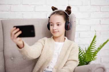 Teknoloji ve internet konsepti - koltukta oturan, müzik dinleyen veya modern akıllı telefonuyla selfie çeken tatlı bir kızın portresi