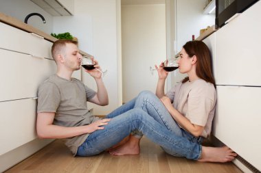 Ev, aşk, flört ve romantizm konsepti. Genç aşık bir çift yerde oturuyor, bir şeyler kutluyor ve parlak mutfakta kırmızı şarap içiyor.