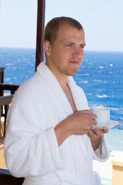 Happy man in bathrobe with cup of coffee standing on balcony — Stock Photo, Image