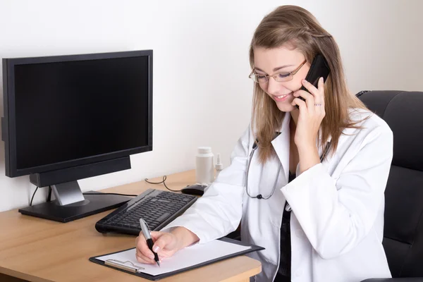 Woman doctor talking by phone and writing something in her offic — Stock Photo, Image