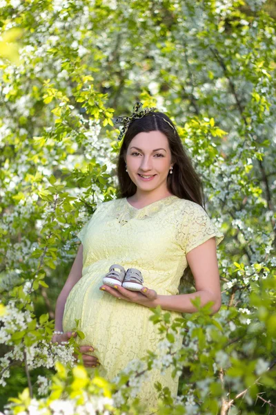 Schwangere Frau mit kleine Schuhe in blühende Kirsche Garten — Stockfoto