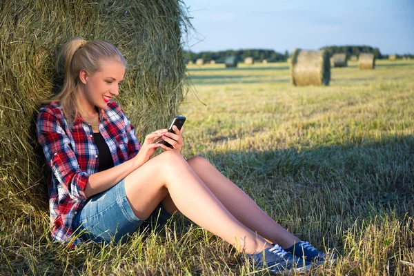 Beautiful blonde woman sitting near haystack in field with mobil — Stock Photo, Image