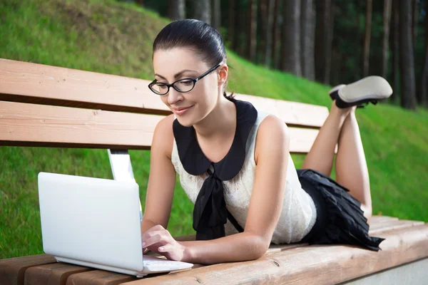 Chica en uniforme escolar usando laptop acostado sobre un banco en el Parque —  Fotos de Stock