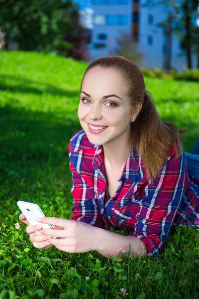 Mosolygó tizenéves lány feküdt az okos telefon a füves Park — Stock Fotó