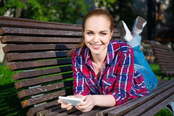 Happy tonårig flicka liggande med smart telefon på bänk i parken — Stockfoto