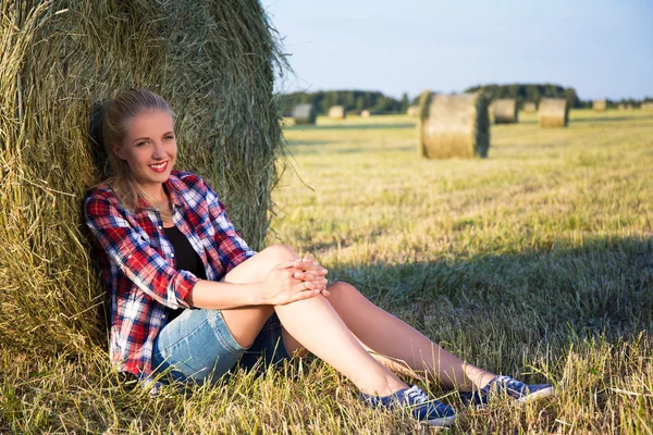 Beautiful blonde woman sitting near haystack in field — Stock Photo, Image