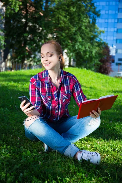 Teenage girl with smart phone and book in park – stockfoto