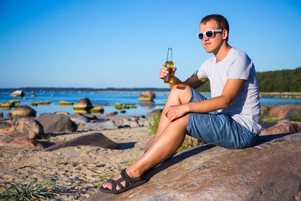 Portrait of handsome man drinking beer on stony beach — Stock Photo, Image