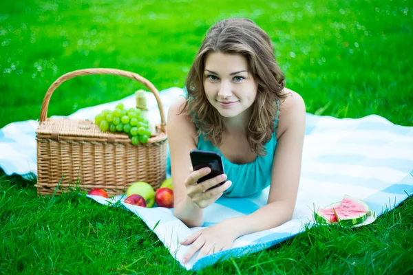 Cute woman with picnic basket and fruits using smart phone in pa — Stock Photo, Image