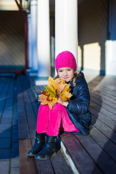 Χαριτωμένο μικρό κορίτσι σε ζεστά ρούχα με κίτρινα φύλλα που κάθεται στο t — Φωτογραφία Αρχείου