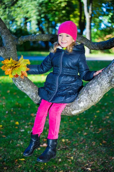 Cute happy girl sitting on tree branch in autumn park — Stock Photo, Image