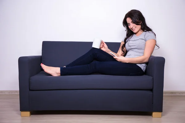 Feliz mulher sentada no sofá com telefone e caneca de chá — Fotografia de Stock