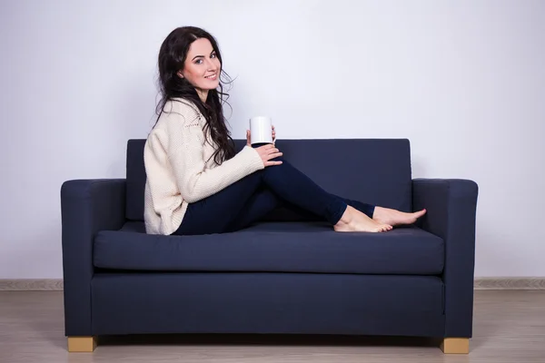 Ung kvinna sitter på soffan med mugg te eller kaffe — Stockfoto