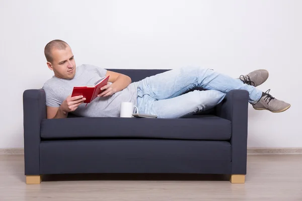 Pohledný muž leží na pohovce a čtení knih s šálkem čaje — Stock fotografie