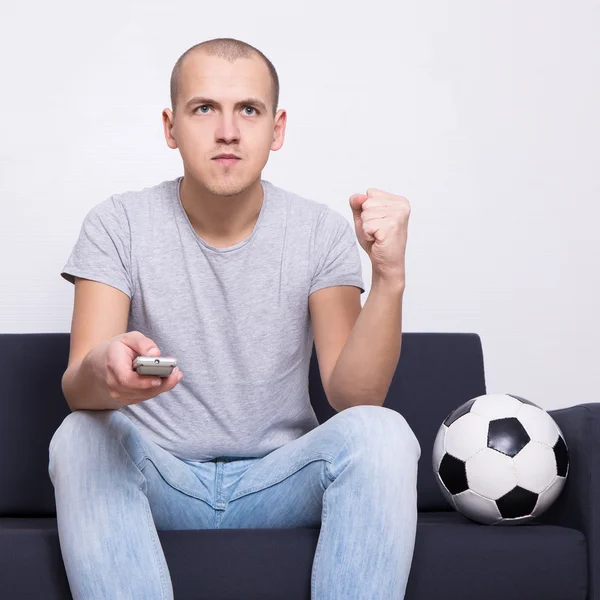 Fan piłki nożnej człowiek siedzi na kanapie z piłką i oglądania telewizji w domu — Zdjęcie stockowe