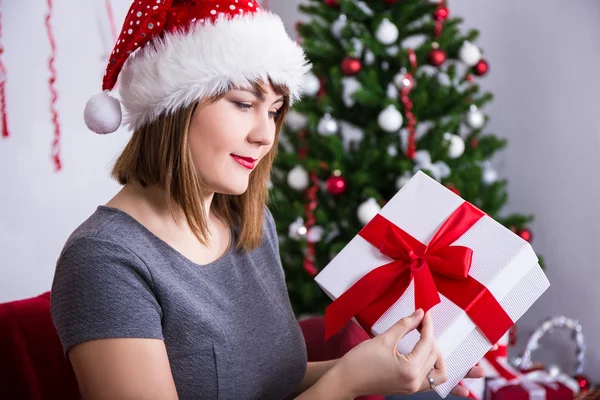 Gelukkig jonge vrouw in Kerstman hoed openen van de doos van de gift in de buurt van Kerstmis t — Stockfoto